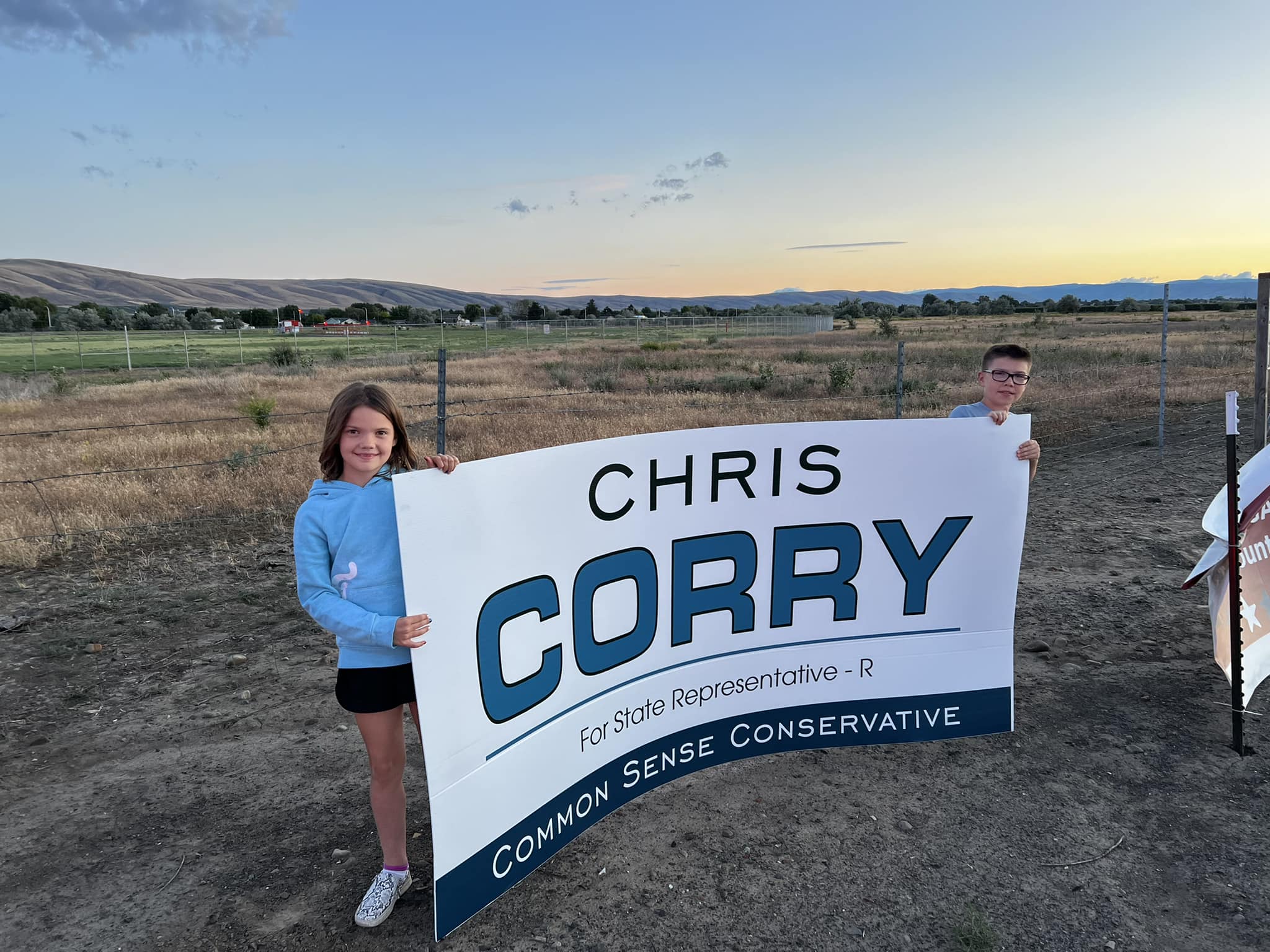 Two of Chris Corry's children holding a large 'Chris Corry — Common Sense Conservative' yard sign at golden hour, with the Yakima Valley landscape behind them
