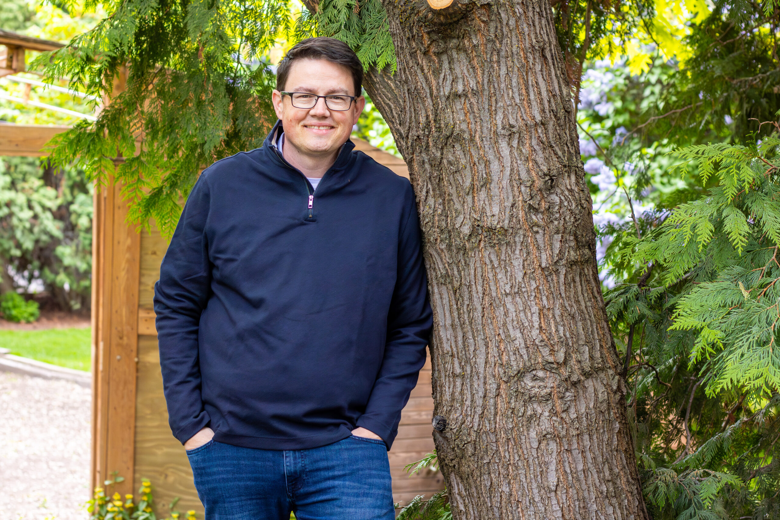 Chris Corry leaning casually against a cedar tree, in a navy quarter-zip pullover