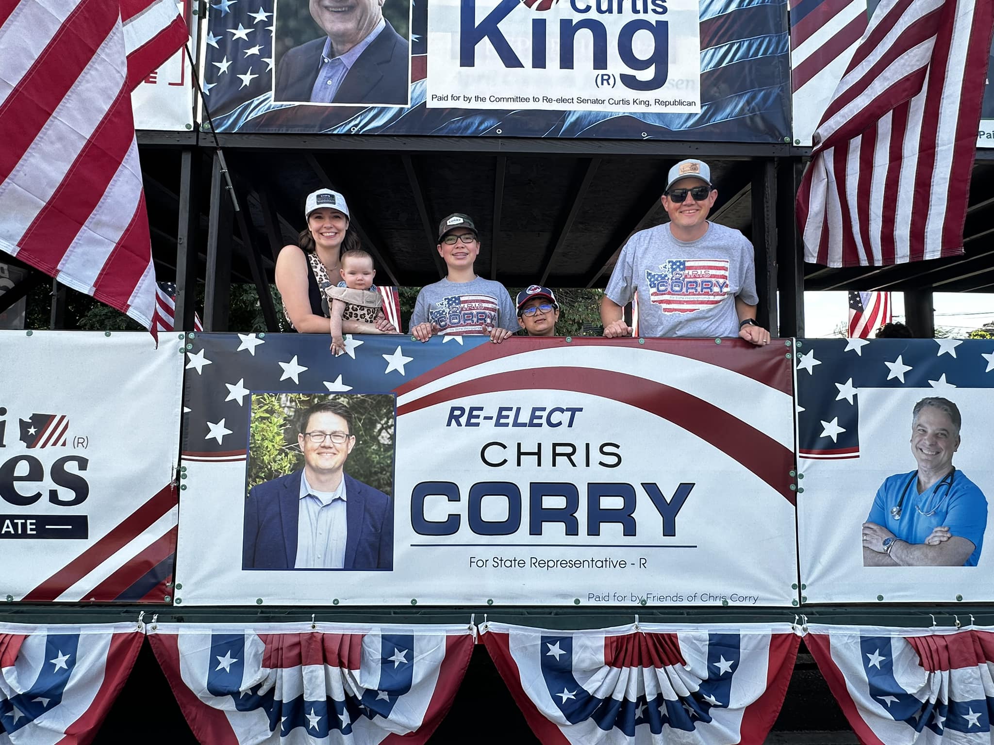 Chris Corry, his wife, and their children on a Yakima Valley parade float, with a 'Re-elect Chris Corry' banner visible and American flags surrounding the float