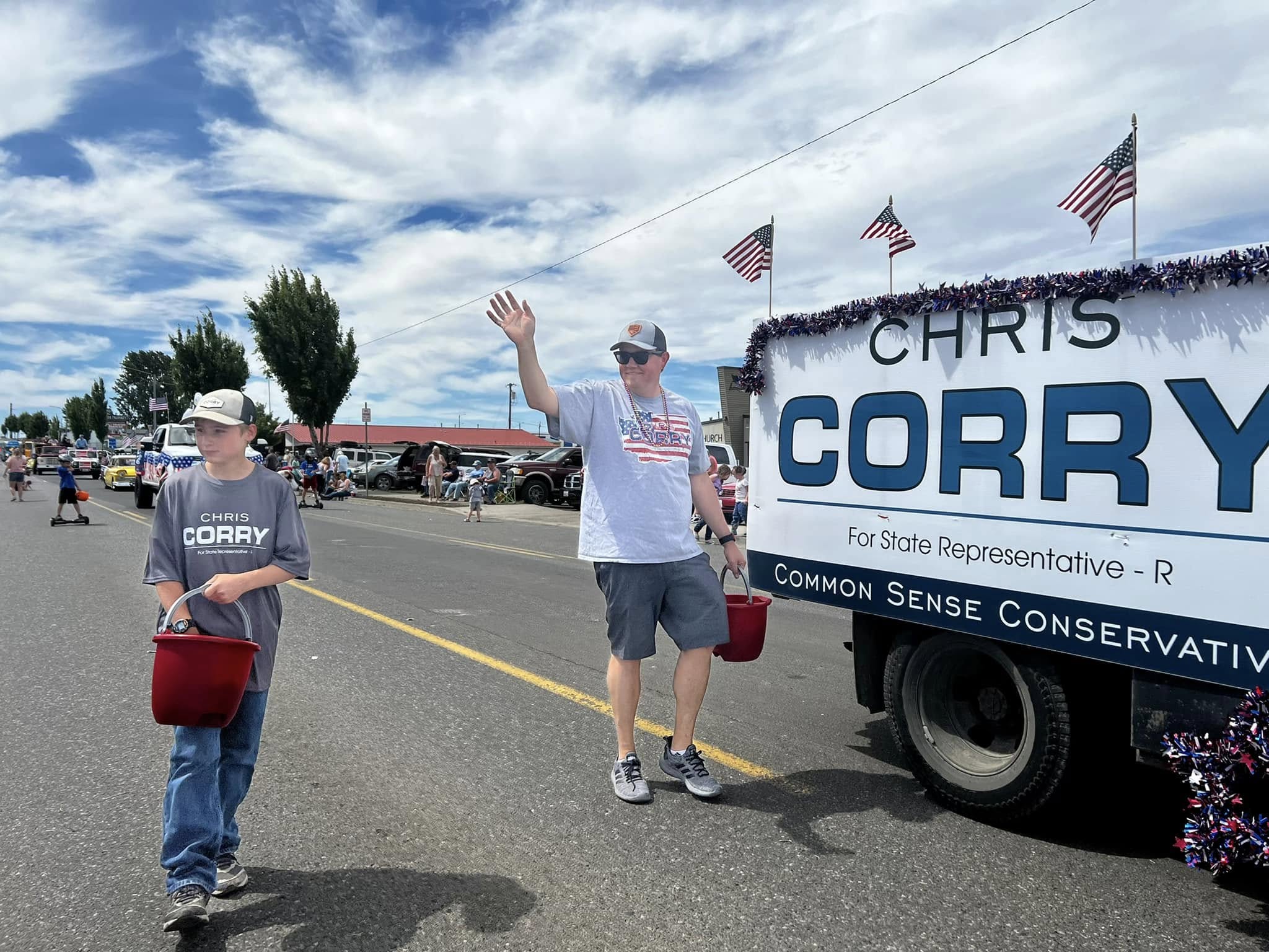 Chris Corry waving in a Yakima Valley parade alongside his Corry campaign float, with American flags and his son walking next to him