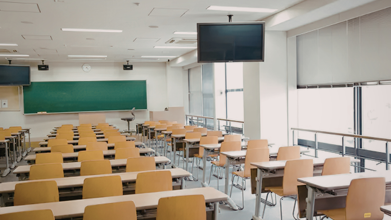 An empty modern classroom with rows of student desks, a green chalkboard at the front, and natural light through wide windows